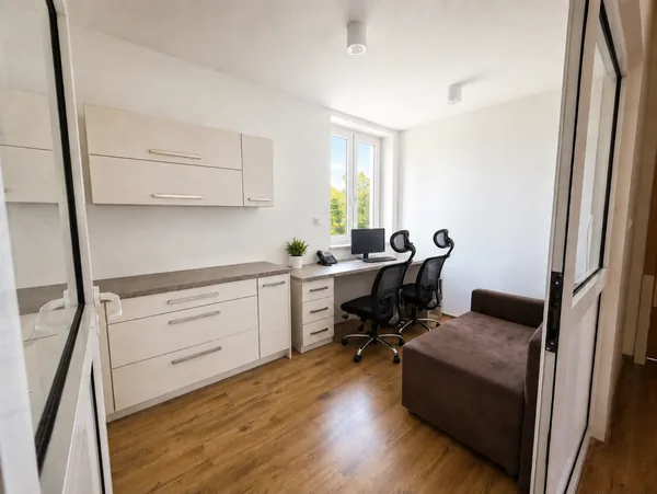 Dyżurka pielęgniarek widziana przez przeszklone drzwi: zabudowa z szafkami, biurko pod oknem z monitorem i telefonem, dwa krzesła biurowe i sofa.