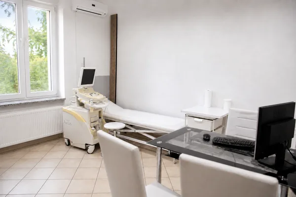 Jasny gabinet USG: aparat USG, kozetka, białe krzesła i szklane biurko z monitorem, za oknem zieleń.
